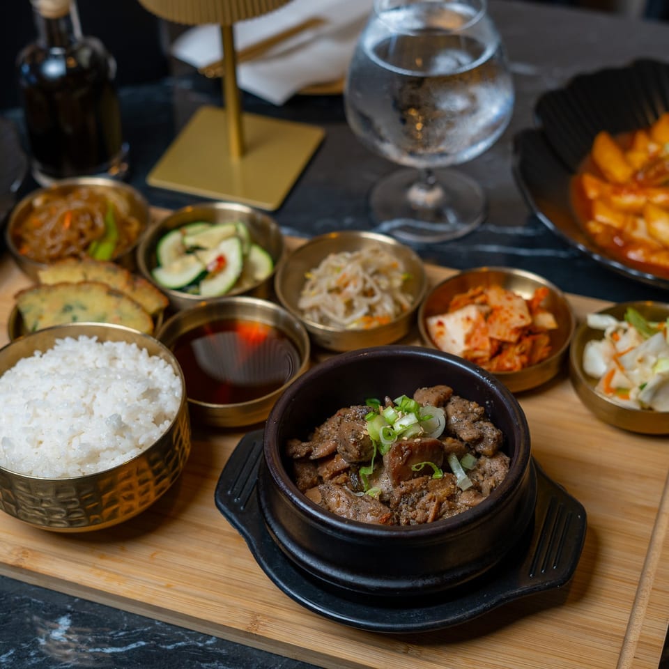 Korean meal spread featuring rice, sizzling beef, kimchi, side dishes, and spicy rice cakes on a wooden serving board.