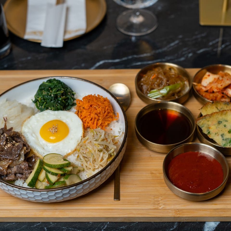 Korean bibimbap with beef, vegetables, fried egg, kimchi, savory pancakes, and dipping sauces on a wooden serving board.
