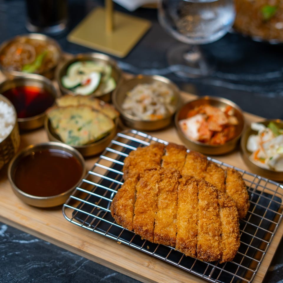 Korean-style breaded cutlet served with white rice, dipping sauces, and assorted side dishes on a wooden platter.