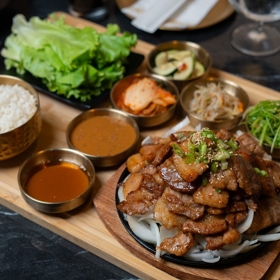 Korean barbecue platter with grilled meat, white rice, lettuce, dipping sauces, kimchi, pickled vegetables, and greens.